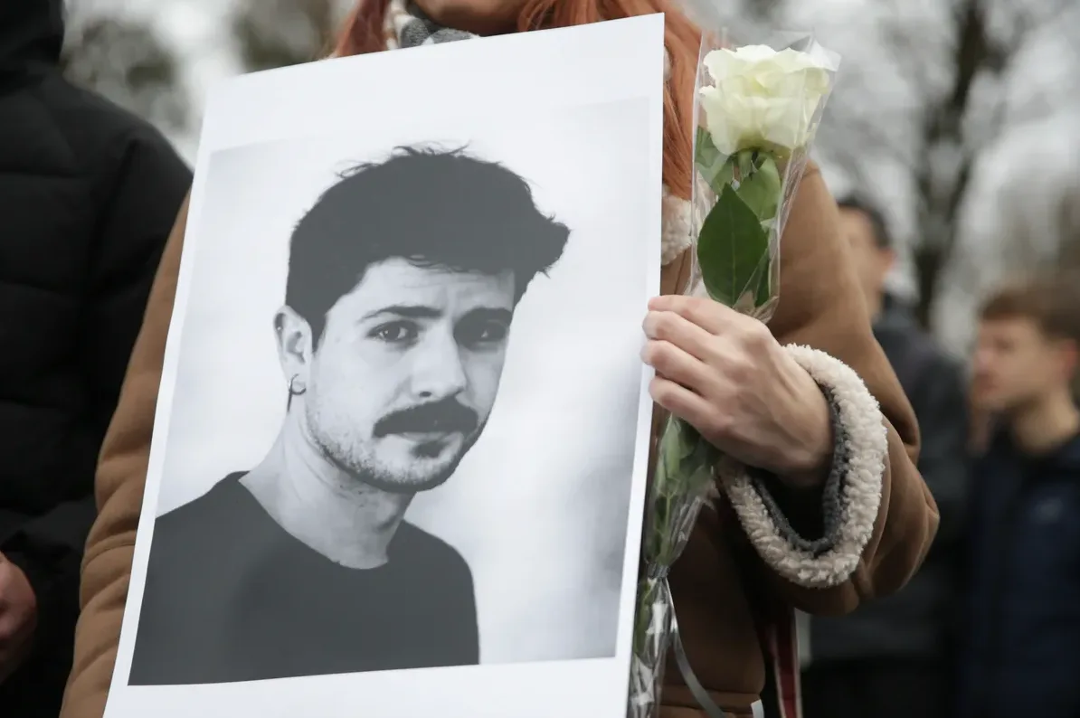 Crno-bijeli portret mladića s brkovima i minđušom u uhu, kojeg drži osoba sa bijelom ružom u ruci.