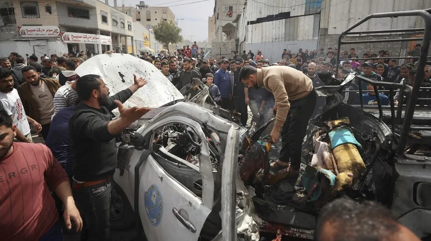 Grupa ljudi okružuje teško oštećeno bijelo vozilo, vjerovatno policijsko, koje je pretrpjelo značajnu štetu, vjerovatno uslijed eksplozije ili napada. Nekoliko ljudi, uključujući jednog muškarca koji gestikulira rukama, djeluju uznemireno ili bijesno, dok drugi stoje i promatraju uništeno vozilo. U pozadini se naziru građevine i veći broj okupljenih civila.