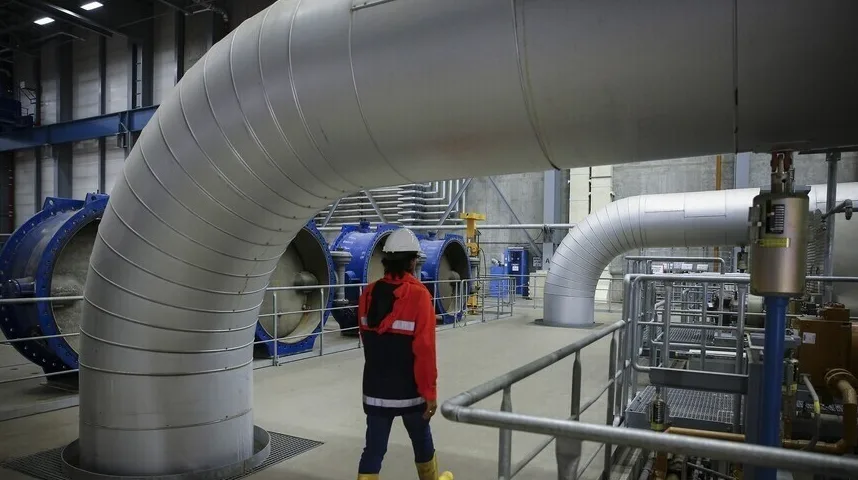 Radnica u bijeloj kacigi i zaštitnom prsluku šeta kroz industrijsko postrojenje s velikim, srebrnim cijevima i plavim ventilatorima u pozadini.