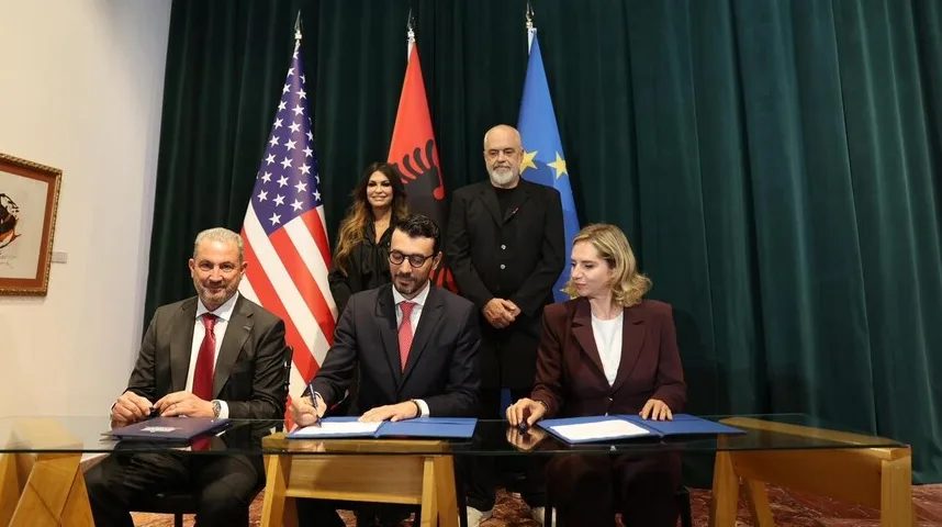 Dvojica muškaraca u odijelima potpisuju dokument na stolu, ispred njih su zastave Sjedinjenih Američkih Država, Albanije i Evropske unije. Pored njih stoje žena i muškarac.