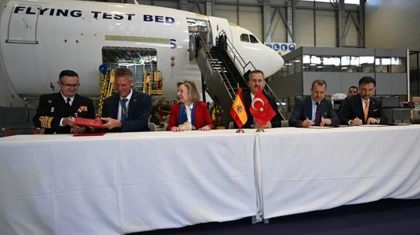Zvaničnici u svečanim odijelima i vojnim uniformama potpisuju dokumente za stolom, sa španskom i turskom zastavom ispred njih, u hangaru pored aviona koji je označen kao "FLYING TEST BED".