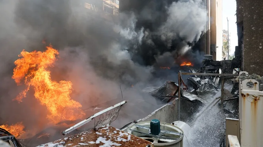 Plameni jezici vatre šire se iz zapaljene zgrade, okruženi gustim crnim dimom. U prvom planu vide se ostaci uništene konstrukcije i opreme, na koju mlazom vode pokušavaju djelovati vatrogasci.