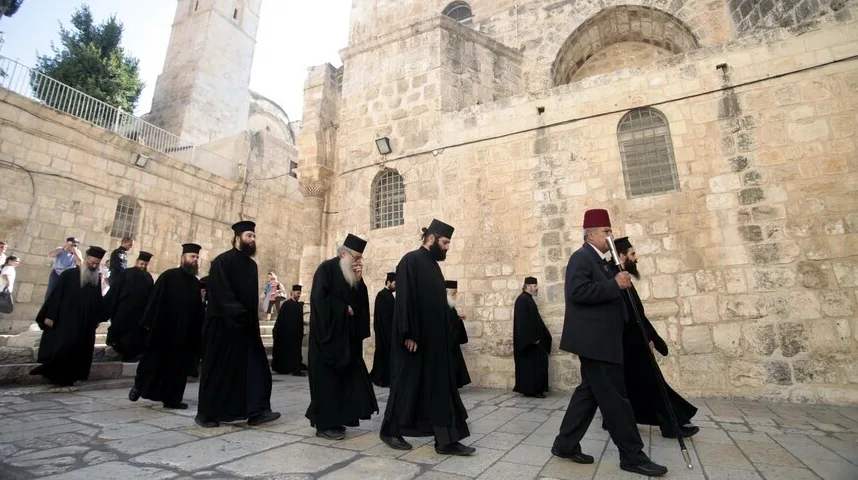 Grupa pravoslavnih sveštenika u crnim haljinama i tradicionalnim kapama, predvođena muškarcem u odijelu sa fesom, maršira kaldrmisanim ulicama Starog grada Jerusalima pored kamenih zdanja.
