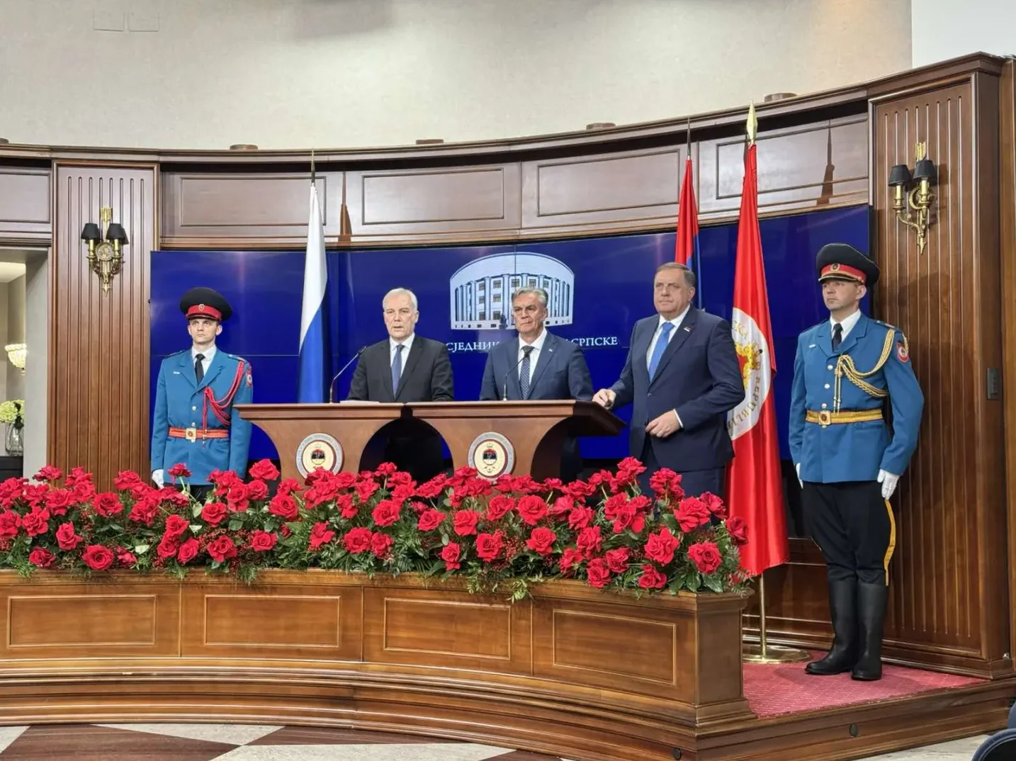 Dva muškarca u svečanim odijelima stoje iza govornice, ukrašene mnoštvom crvenih ruža, dok ih dva pripadnika počasne garde u uniformama posmatraju.