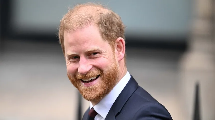 Princ Harry se smije, noseći tamno odijelo i tamnu kravatu.