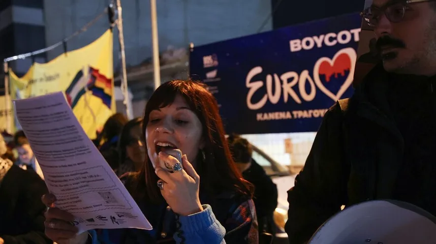 Žena drži papir s tekstom i govori u megafon na protestu, s natpisom "BOYCOT EUROVISION" u pozadini.