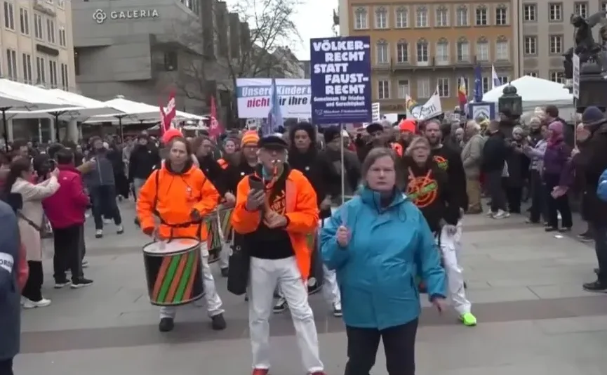 Grupa ljudi maršira ulicom sa bubnjevima i transparentima u rukama.