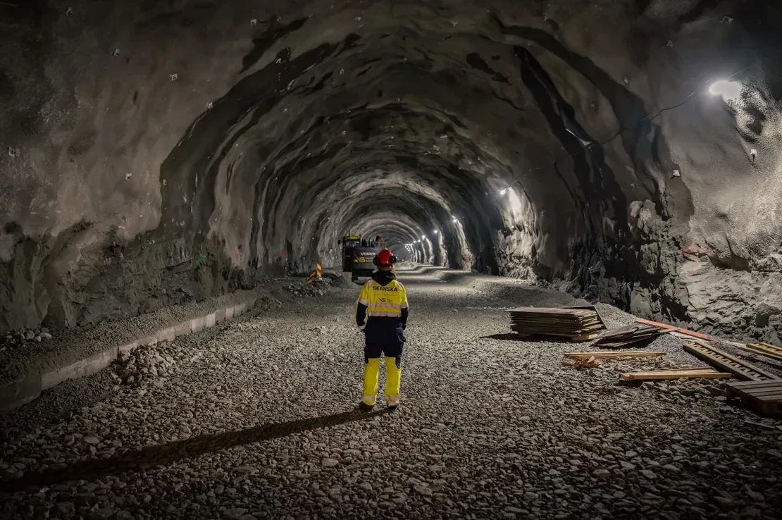 Radnik u zaštitnoj opremi stoji u osvijetljenom tunelu u izgradnji, gledajući prema kraju tunela gdje se nazire mašinerija.
