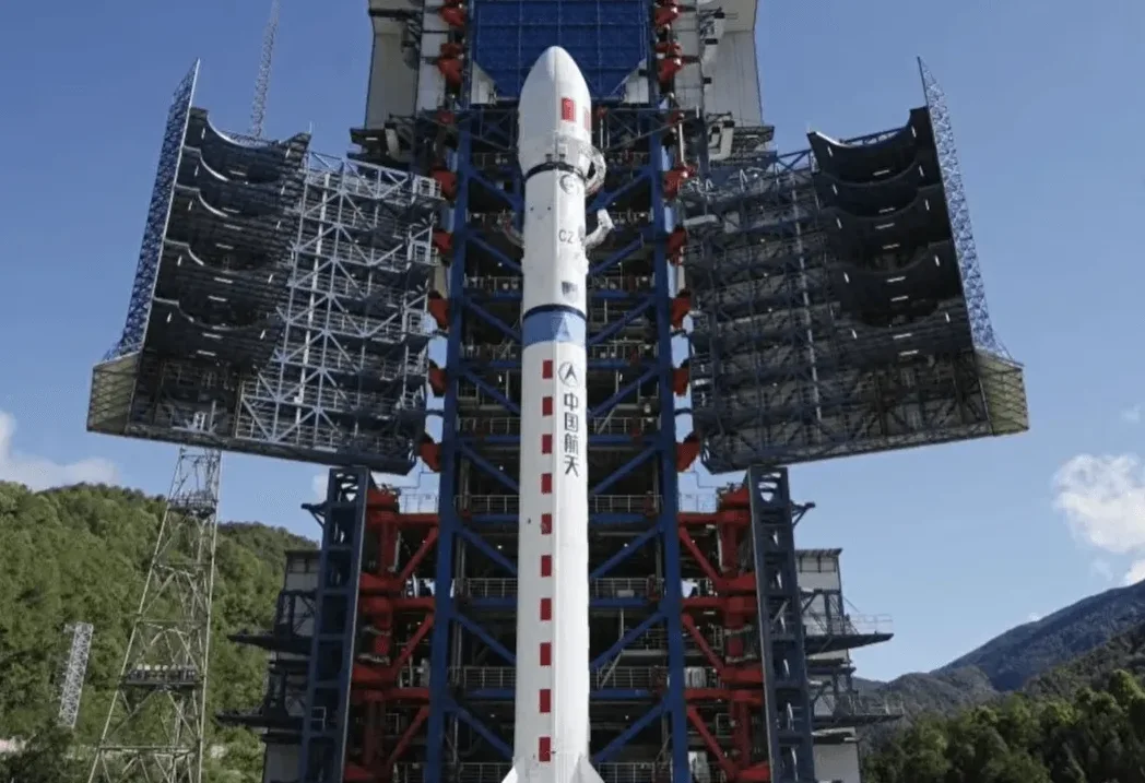 Kineska raketa Čangdžen sa simbolom "Kineski svemir" ispisan kineskim slovima, stoji uspravno na lansirnoj rampi sa otvorenim potpornim krilima.