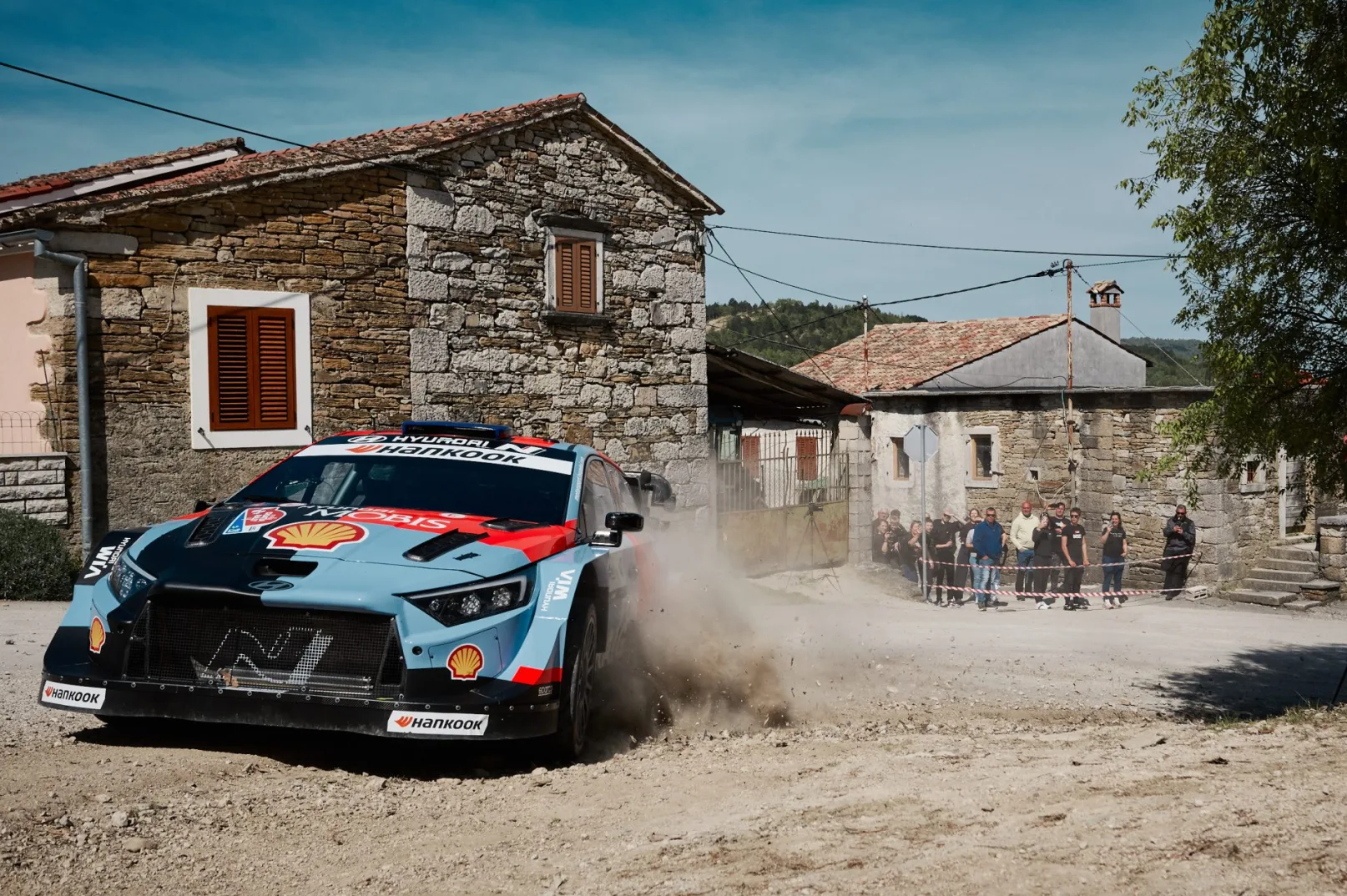 Rally automobil Hyundai u plavoj, crvenoj i crnoj boji, juri kroz prašnjavo seosko naselje s kamenim kućama, dok ga sa strane promatra grupa gledalaca.