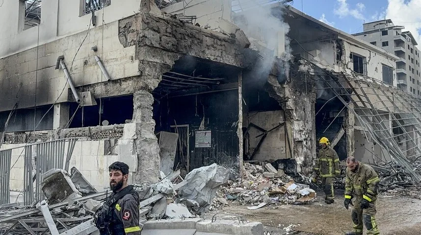 Dva vatrogasca u zaštitnoj opremi pregledaju velika razaranja na zgradi, sa stubovima dima koji se dižu.