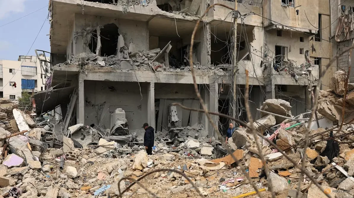 Razrušena stambena zgrada sa vidljivim oštećenjima od bombardovanja, sa ruševinama na prvom planu i nekoliko ljudi koji pregledaju štetu.