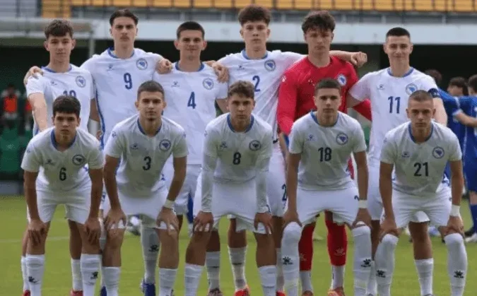 Mlada nogometna reprezentacija Bosne i Hercegovine u bijelim dresovima stoji na nogometnom terenu, spremni za utakmicu.