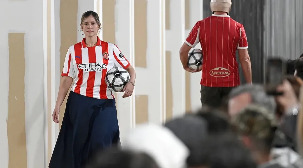 Žena nosi crveno-bijeli nogometni dres s natpisom "ETIHAD AIRWAYS" i tamnoplavu suknju, noseći nogometnu loptu.