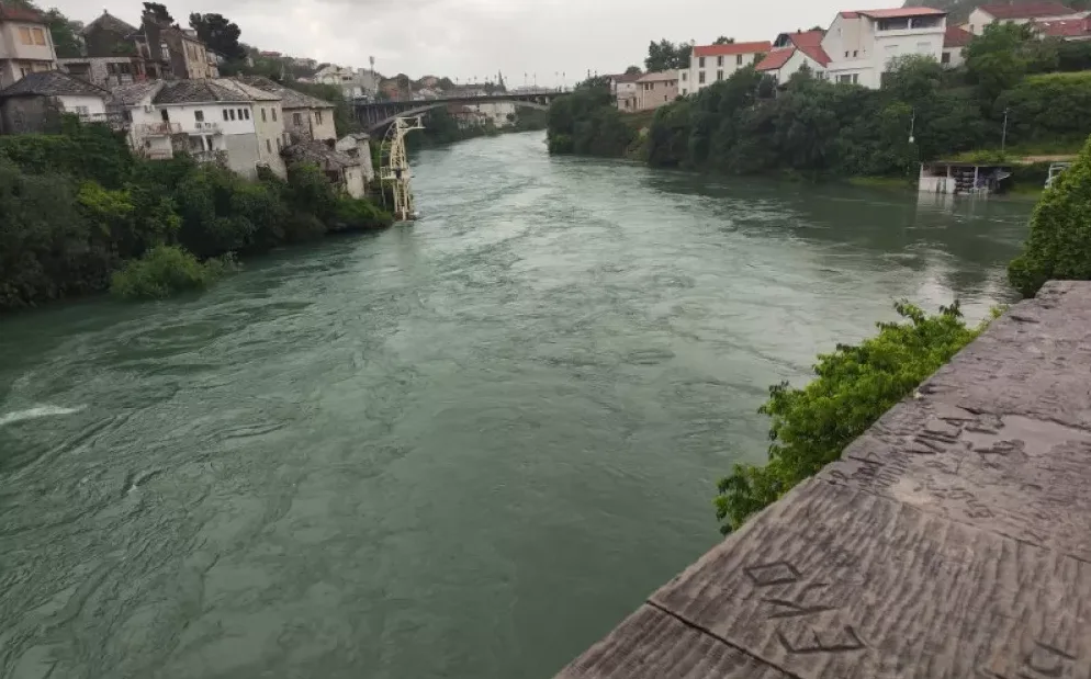 Rijeka Neretva sa lijeve obale, sa starim kućama i drvećem, te mostom u daljini.