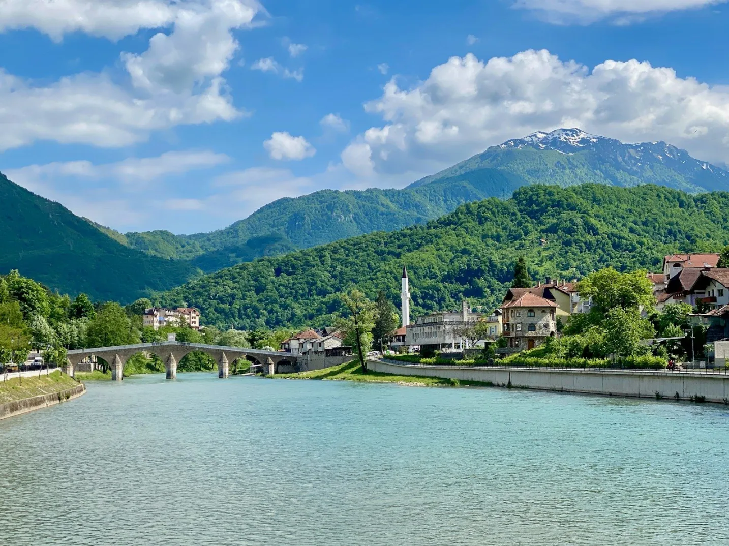 Pogled na rijeku sa kamenim mostom, gradom i zelenim planinama u pozadini.