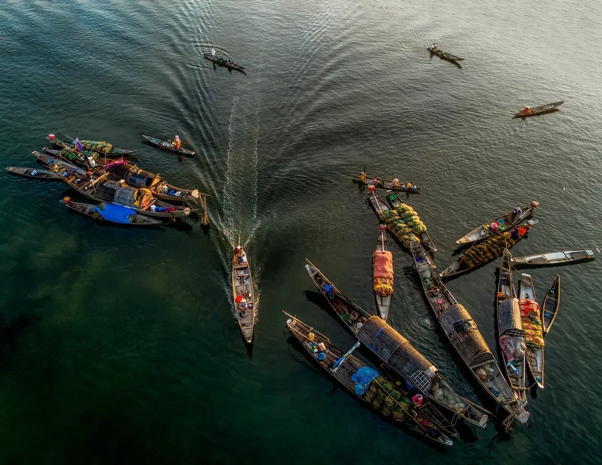 Pogled iz zraka na mnoštvo ribarskih čamaca natovarenih robom na tamnoj, mirnoj rijeci, koji plove u različitim smjerovima.
