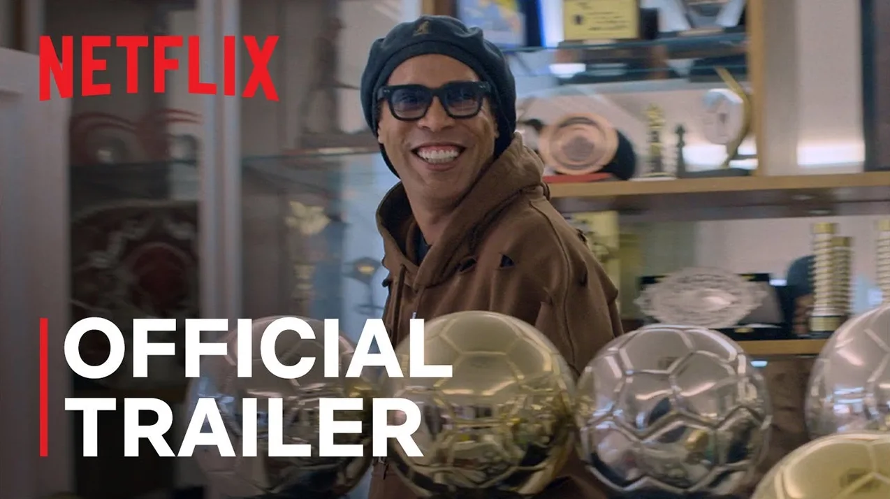 Ronaldinho u smeđoj dukserici i crnoj beretki, sa naočalama, smiješi se pored zlatnih i srebrnih fudbalskih lopti i trofeja. "OFFICIAL TRAILER" i "NETFLIX" natpisi su vidljivi.