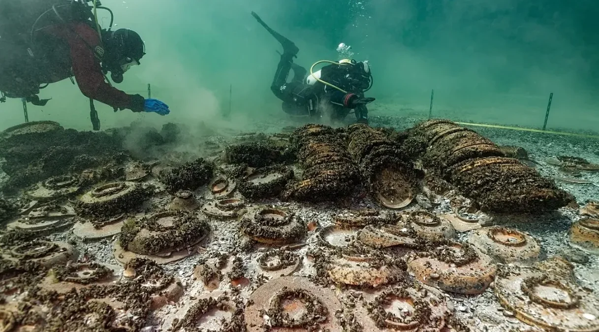 Ronilac u crvenom odijelu pažljivo pregleda ostatke antičkih amfora na morskom dnu, dok drugi ronilac radi u pozadini.