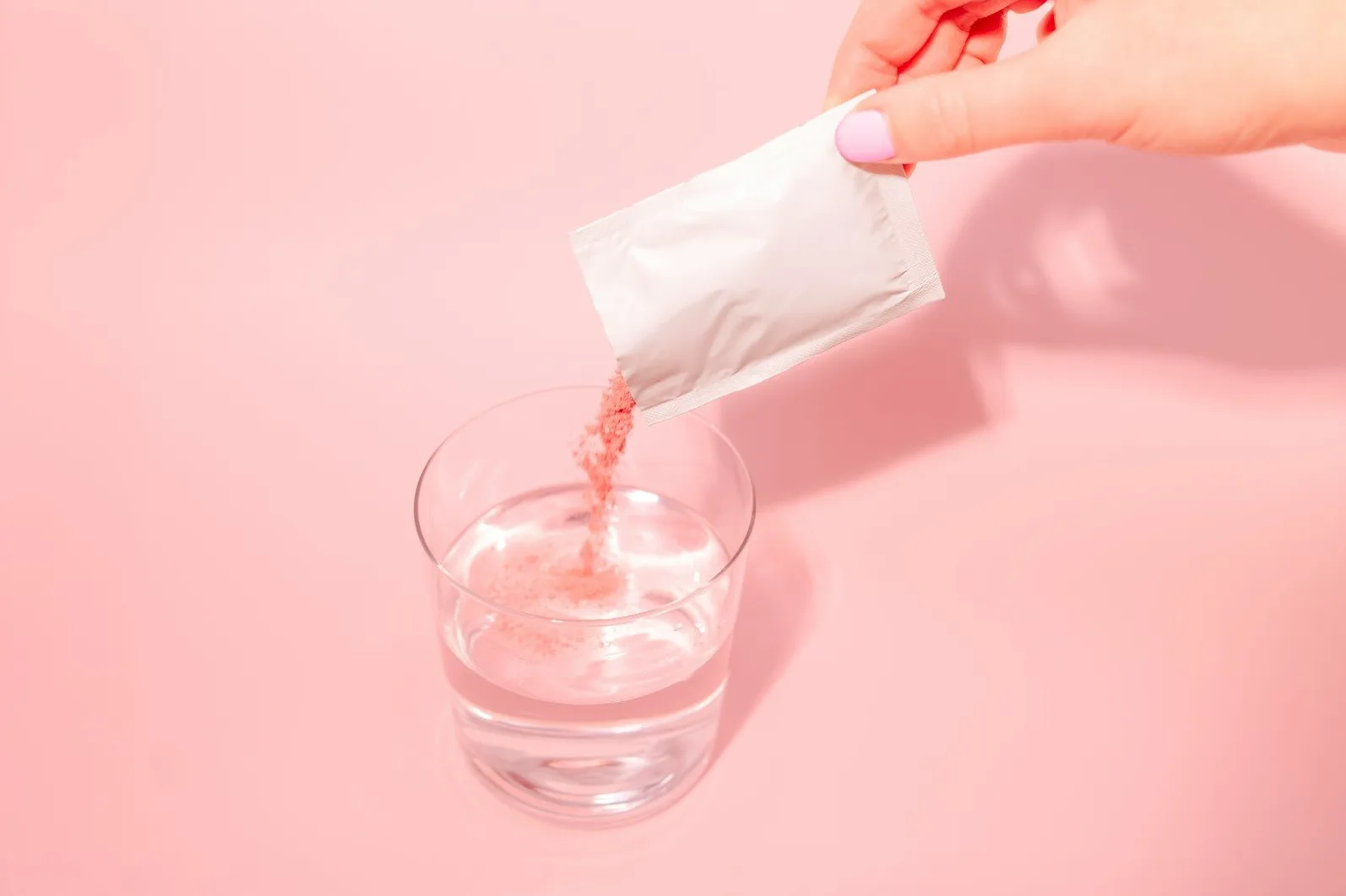 Ruka s urednim noktima drži mali bijeli paketić iz kojeg se prosipa rozi prah u čašu prozirne vode na ružičastoj pozadini.