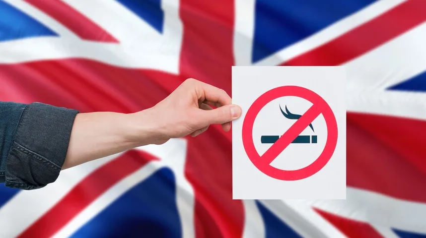 Znak "zabranjeno pušenje" u crvenom krugu s precrtanom cigaretom, koji drži osoba ispred zamućene zastave Ujedinjenog Kraljevstva.