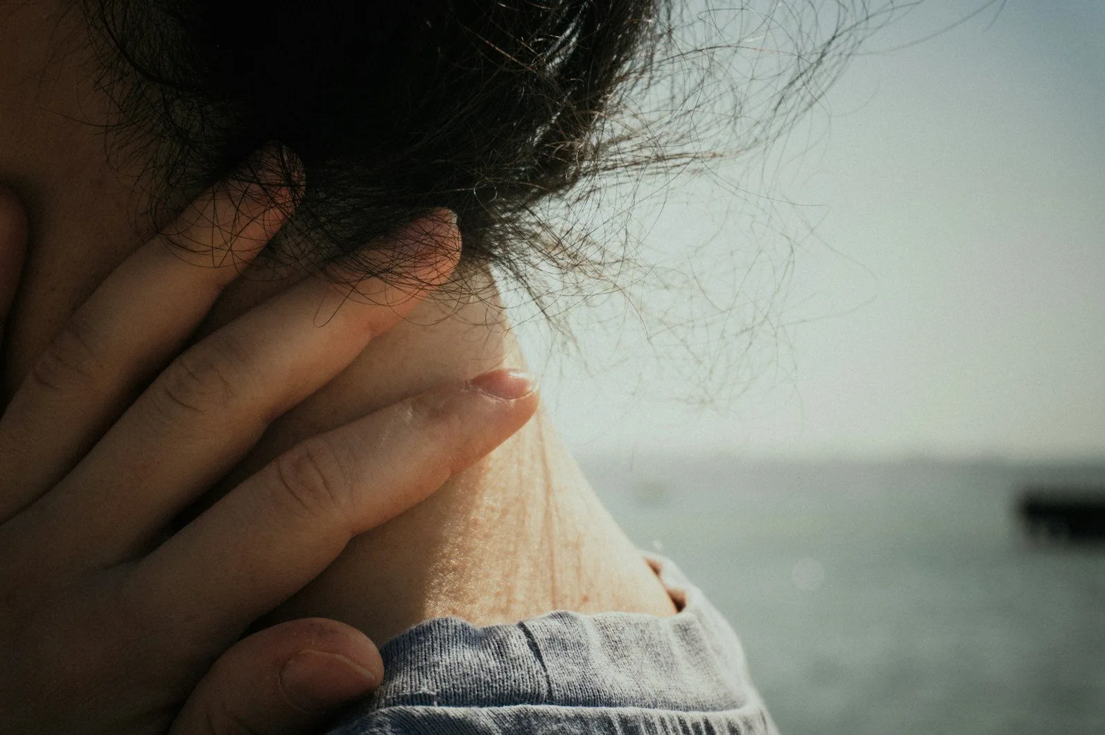 Ruka nježno dodiruje vrat osobe, s kosom koja leprša na vjetru i maglovitim prizorom mora u pozadini.