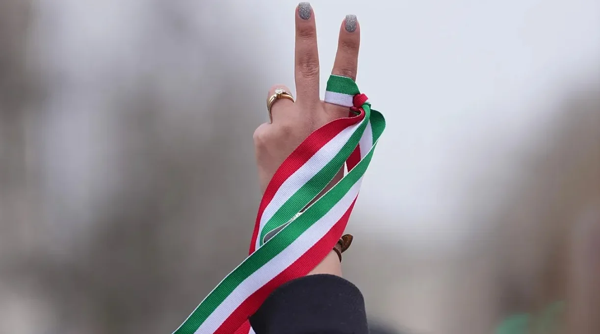Ruka podignuta u znak pobjede, s prstima formiranim u V, okićena iranskom zastavom u obliku trake.