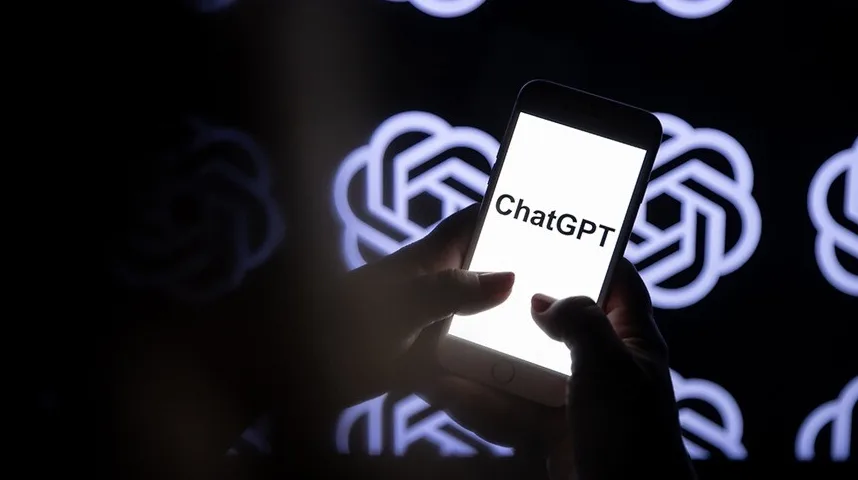 Ruke drže pametni telefon na kojem je prikazan natpis "ChatGPT", s tamnom pozadinom na kojoj se naziru stilizovani logo ChatGPT.