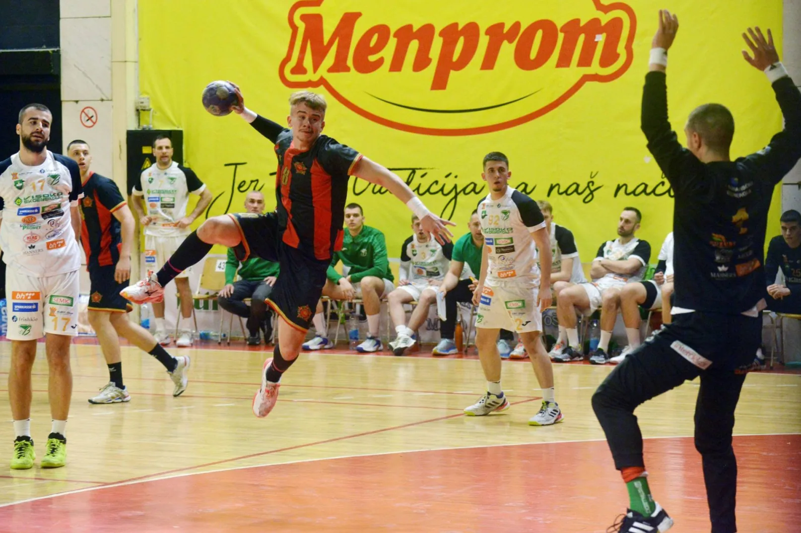 Rukometni igrač u crno-narandžastom dresu skače i drži loptu u ruci, spremajući se da šutira ka golu. Na sebi ima broj 8.