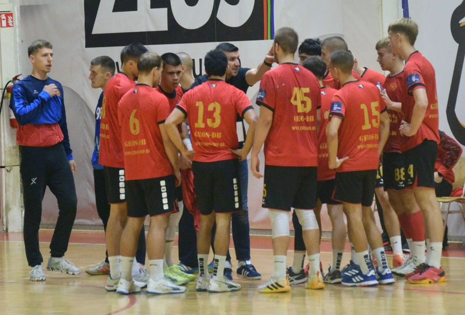 Rukometni tim u crvenim dresovima sa brojevima okupljen na parketu dvorane, dok im trener daje upute.