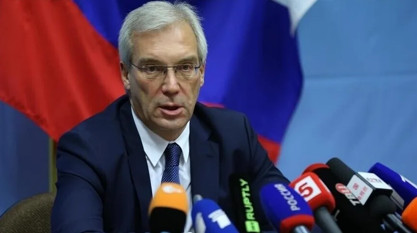 Ruski zvaničnik sa naočarima na glavi, obučen u tamno odijelo i plavu kravatu, govori za govornicom okružen brojnim mikrofonima medijskih kuća, sa zastavom Ruske Federacije u pozadini.