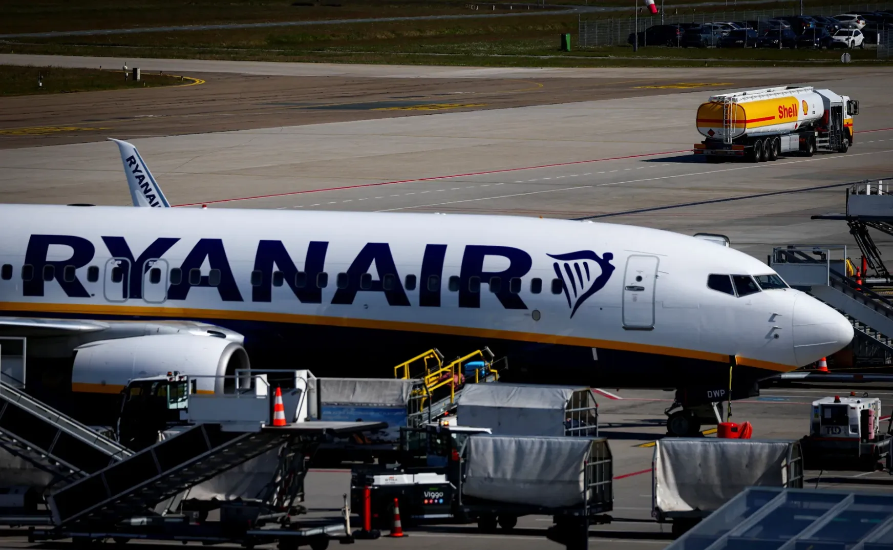 Ryanair avion sa crvenom i žutom trakom na trupu, parkiran na aerodromu. U pozadini se vidi kamion cisterna Shell.
