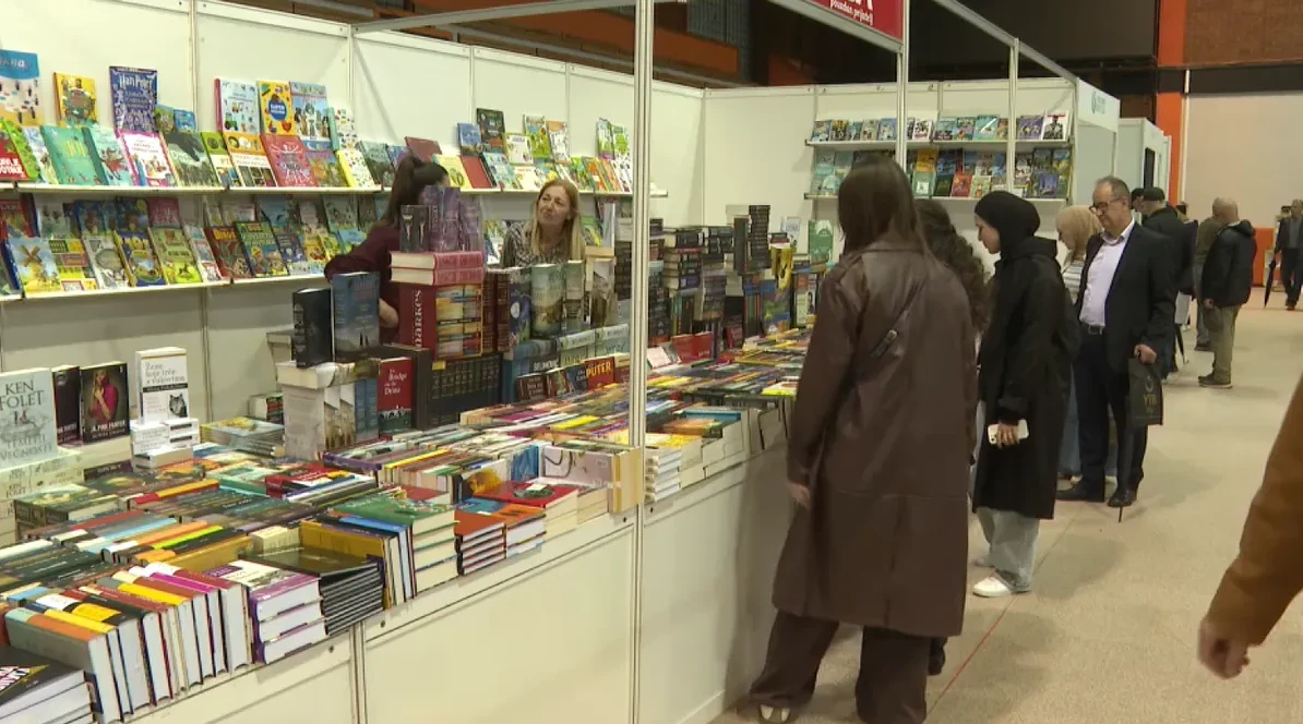 Prodajna tezga puna raznovrsnih knjiga na sajmu knjiga, sa posjetiocima koji razgledaju ponudu.