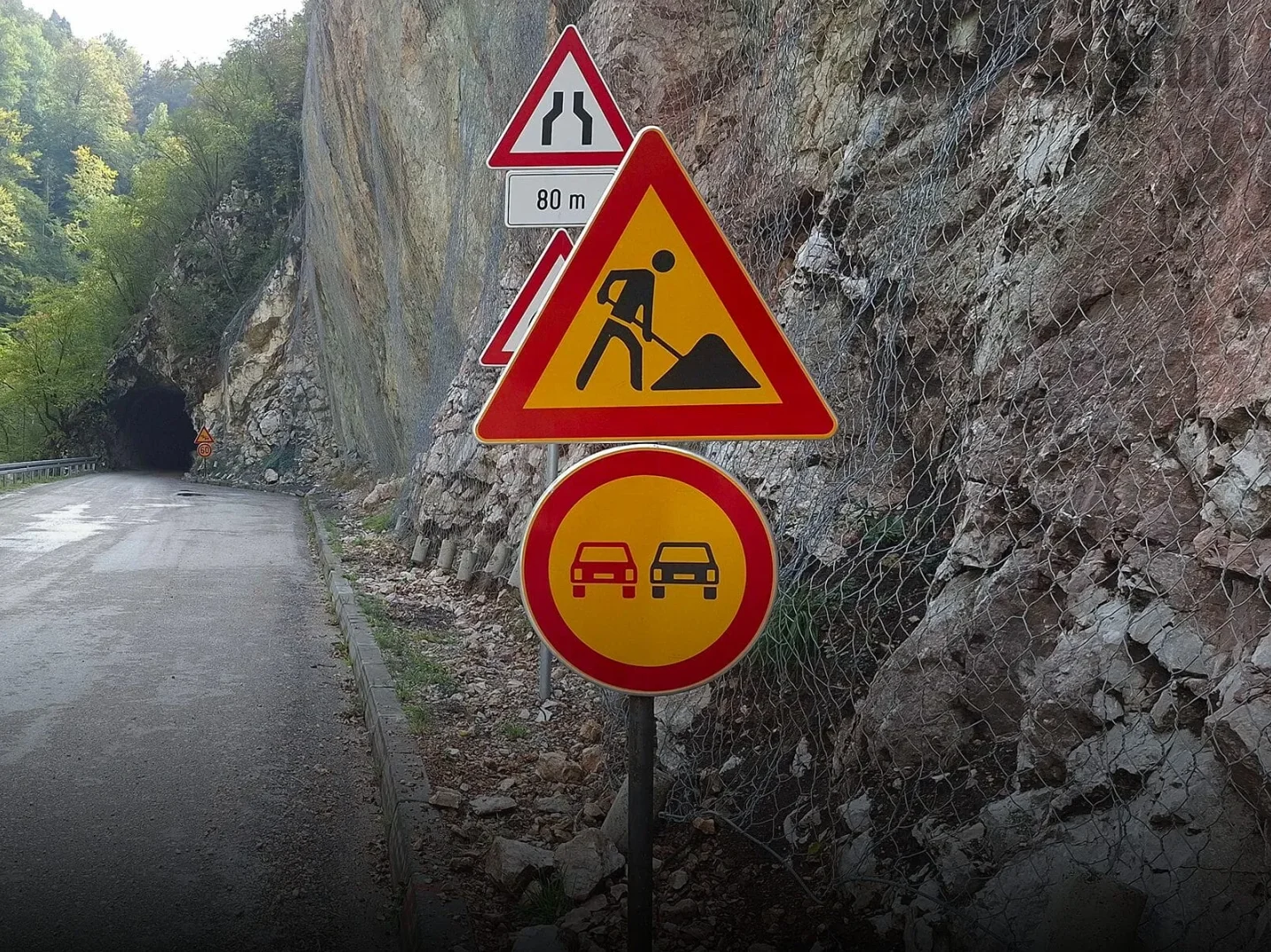 Saobraćajni znaci upozorenja za radove na cesti i zabranu preticanja pored planinskog puta i tunela.