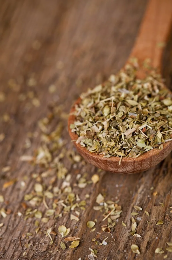 Drvena kašika puna sušenog origana, sa rasutim začinskim biljem po drvenoj površini.