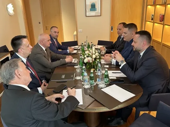 Grupa muškaraca u odijelima sjedi za velikim okruglim stolom na sastanku, na stolu su boce vode, vaze s cvijećem i papiri.