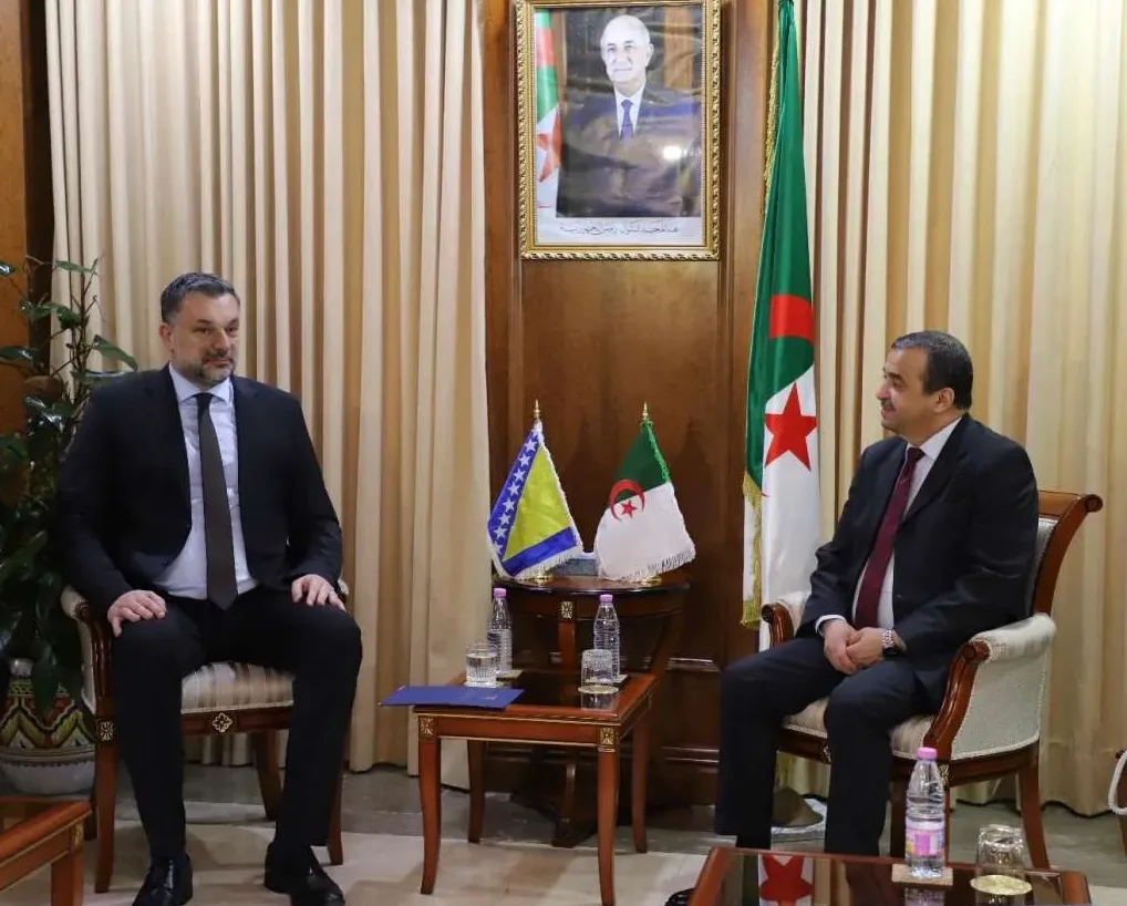 Dvojica muškaraca u odijelima sjede jedan naspram drugoga u uredu, zastave Bosne i Hercegovine i Alžira.