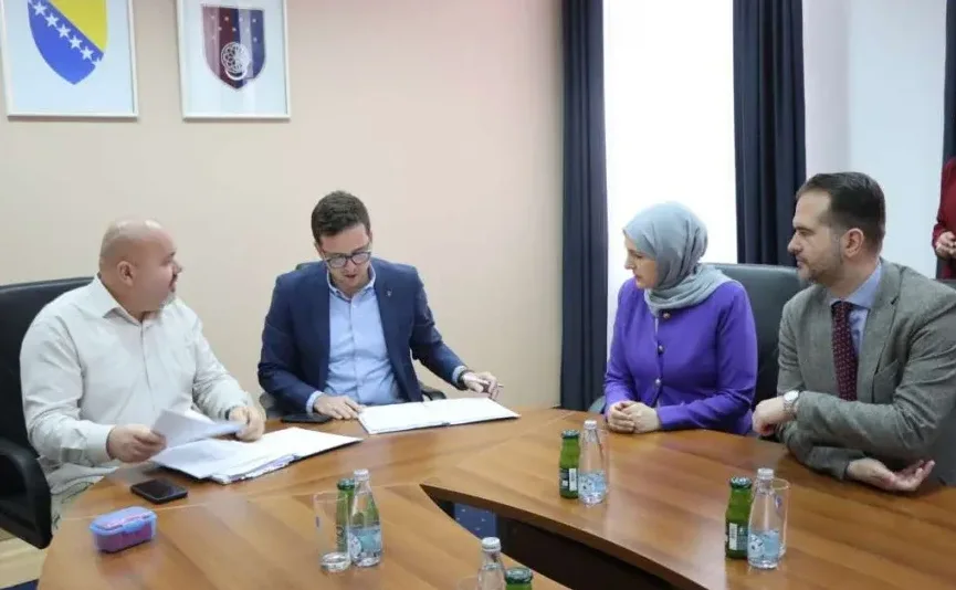 Četiri osobe sjede za okruglim stolom u kabinetu, na stolu su dokumenti, flašice s vodom i sokom. Na zidu iza njih nalaze se zastave Bosne i Hercegovine i nekog drugog entiteta ili institucije.