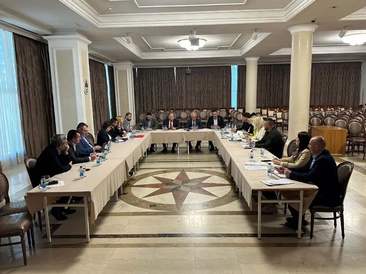 Grupa ljudi sjedi za dugačkim stolom u obliku potkovice tokom sastanka u konferencijskoj sali.