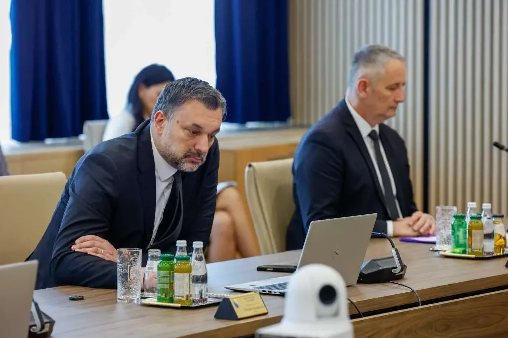 Dva muškarca u odijelima sjede za stolom tokom sastanka, sa laptopom i bocama ispred sebe.