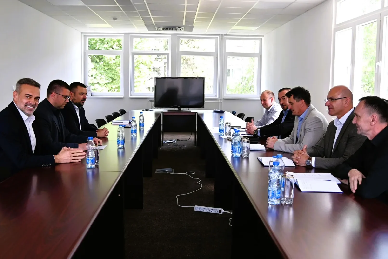 Grupa poslovnih ljudi sjedi za dugim stolom u prostoriji za sastanke, okrenuti jedni drugima.