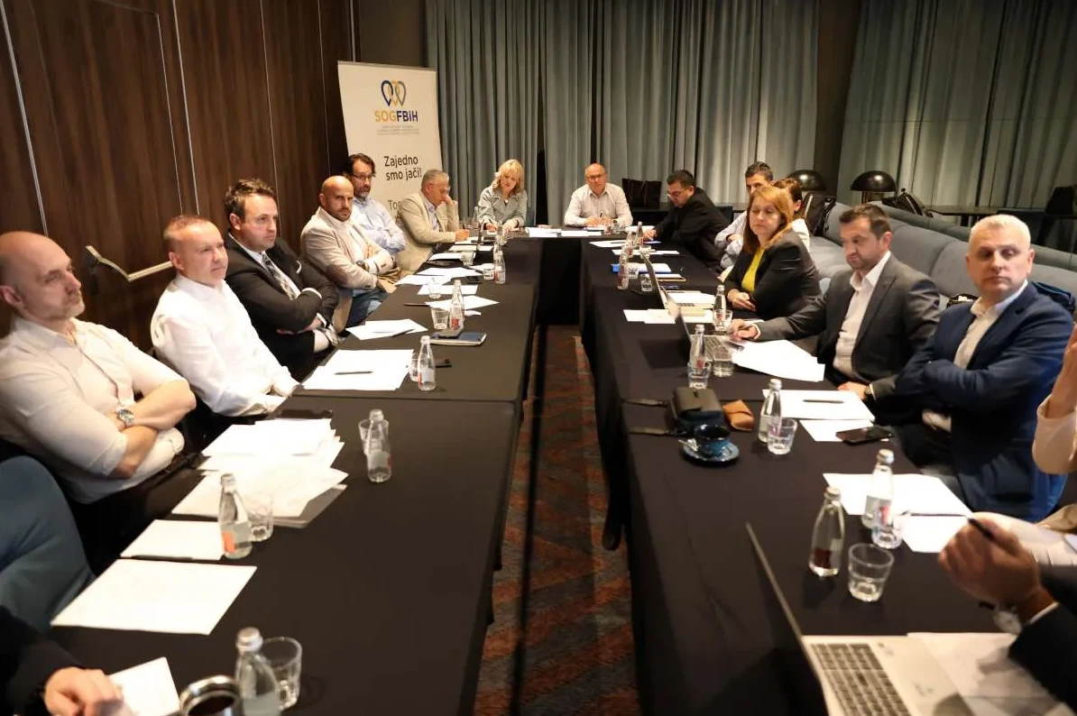 Grupa ljudi sjedi za crnim stolom na sastanku, sa natpisom SOGFBIH u pozadini.