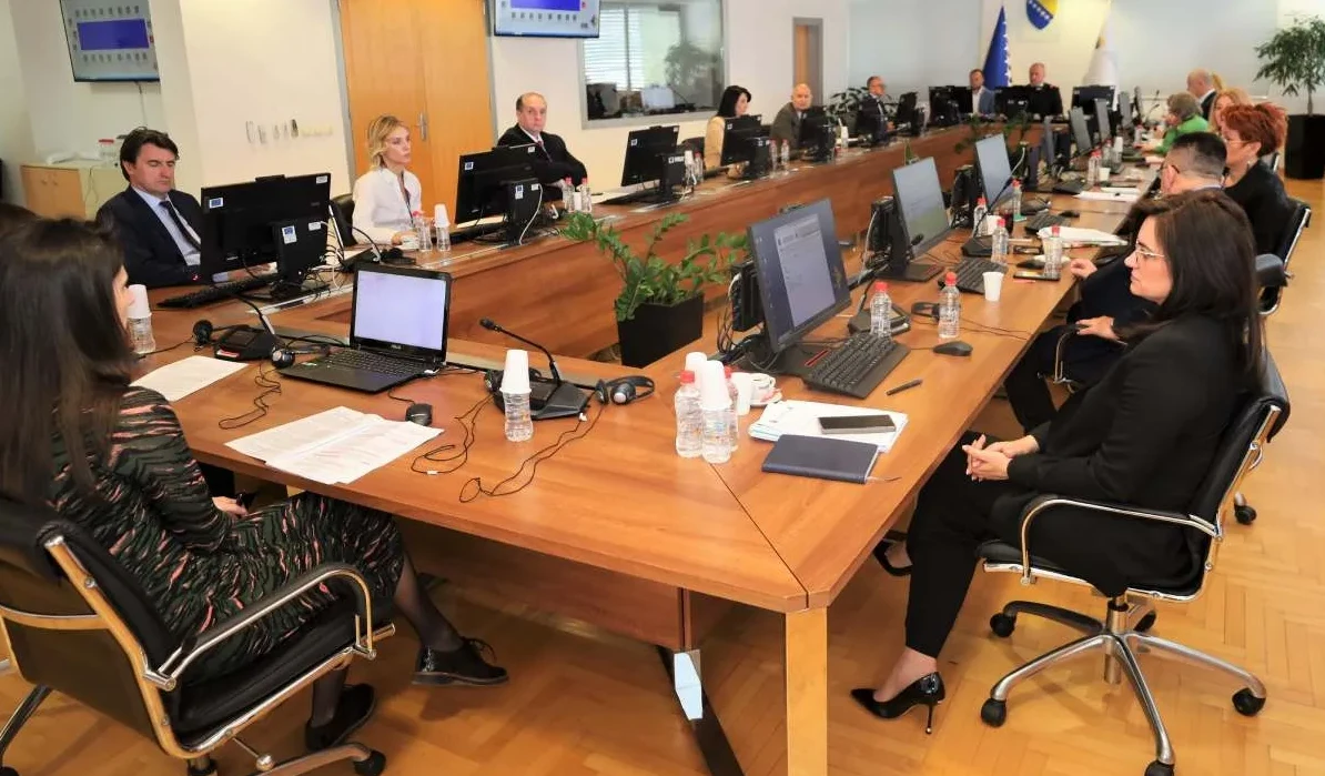 Više osoba sjedi za velikim drvenim stolom u prostoriji nalik na konferencijsku salu, radeći na laptopima i računarima.