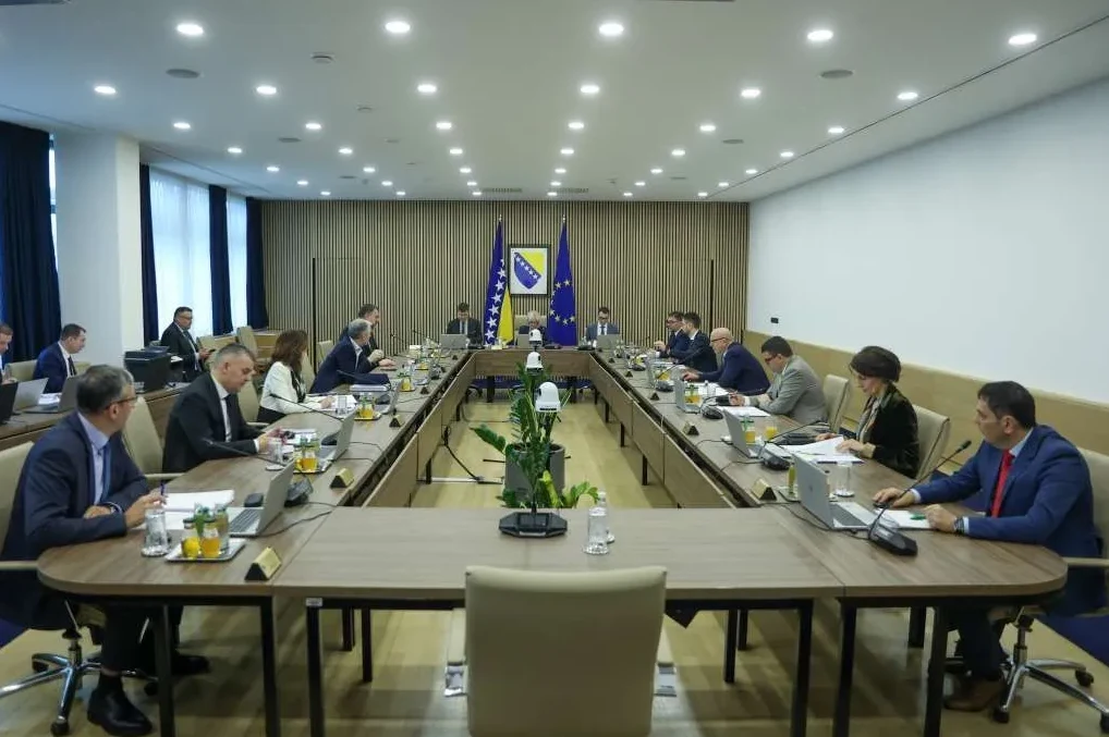 Grupa ljudi u odijelima sjedi za velikim U-oblikovanim stolom na sastanku, okruženi zastavama Bosne i Hercegovine i Evropske Unije.