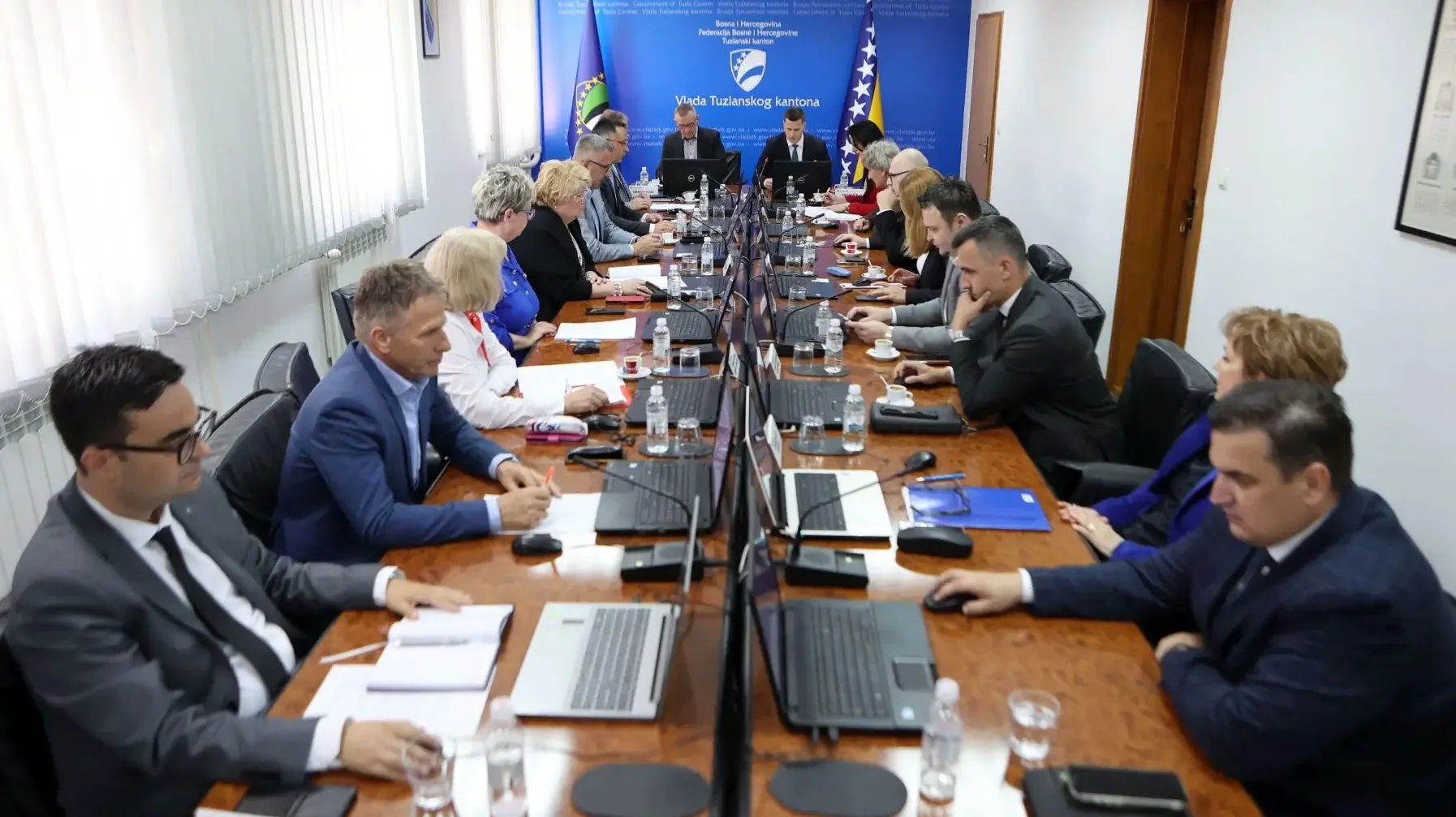 Članovi Vlade Tuzlanskog kantona okupljeni oko dugog drvenog stola, radeći na svojim laptopima i papirima.
