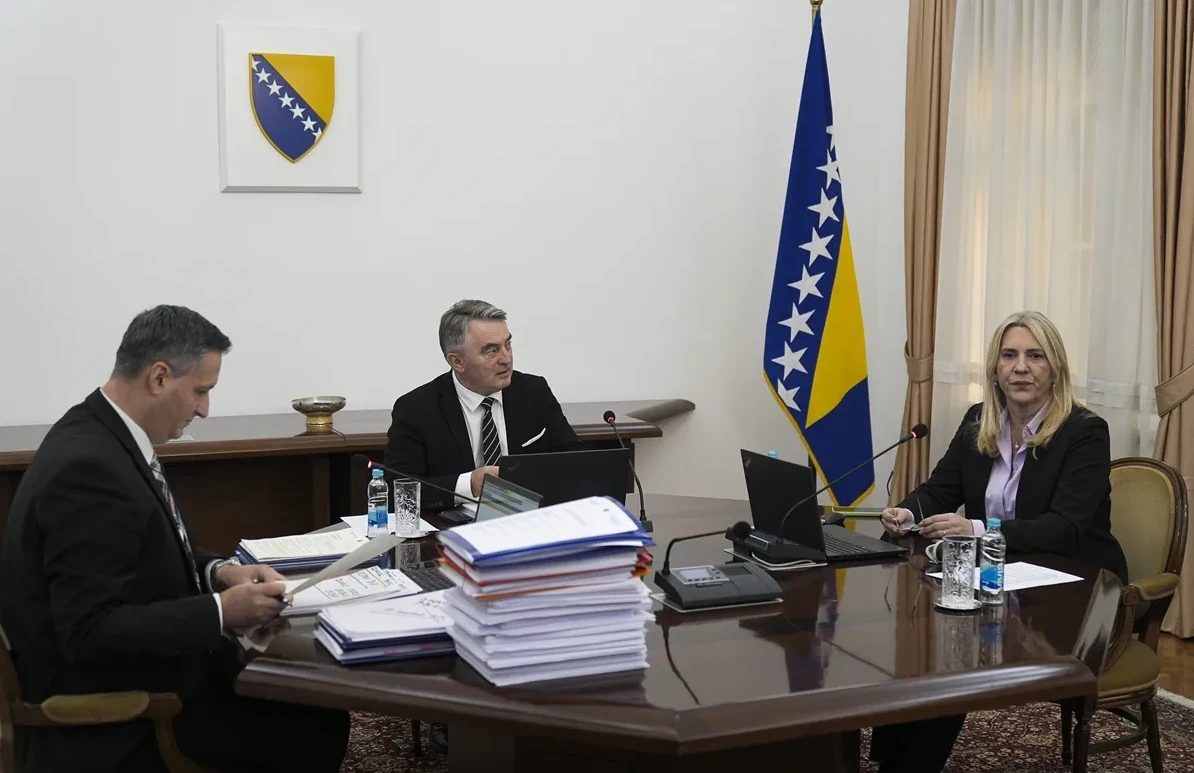Tri zvaničnika Bosne i Hercegovine sjede za velikim drvenim stolom u kancelariji, ispred njih su dokumenti, laptopi i boce vode. Zastava Bosne i Hercegovine i grb nalaze se na zidu u pozadini.