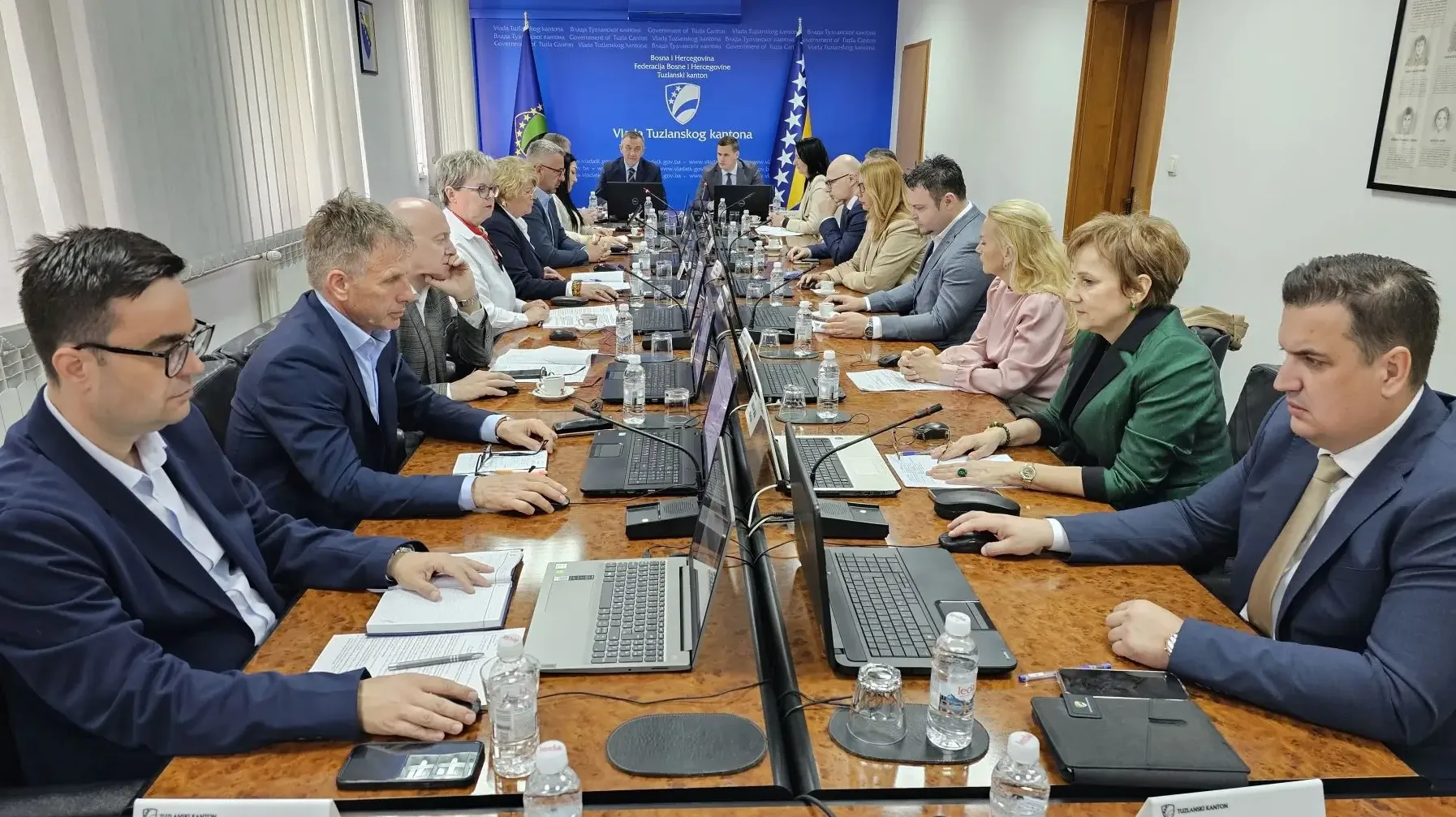 Grupa ljudi na sastanku sjedi oko dugačkog stola, svi nose odijela i gledaju u dokumente ili laptope ispred sebe.