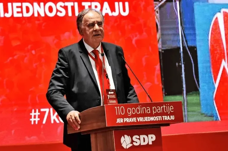 Političar u crnom odijelu i crvenoj kravati govori sa podijuma.