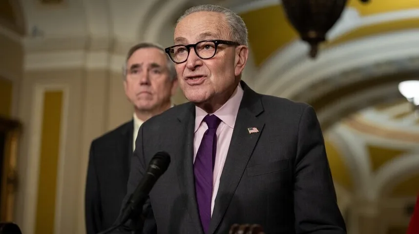 Američki senator Chuck Schumer govori u mikrofon, noseći tamno odijelo, roze košulju i ljubičastu kravatu.