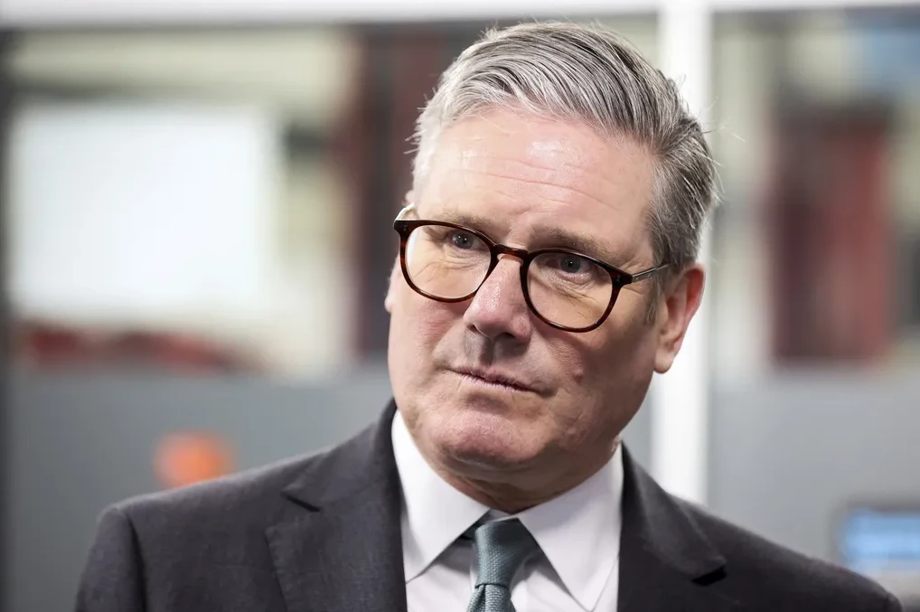 Portret Sir Keira Starmera kako gleda u stranu sa trendovskim smeđim naočalama, odjeven u tamno odijelo i svijetloplavu kravatu.